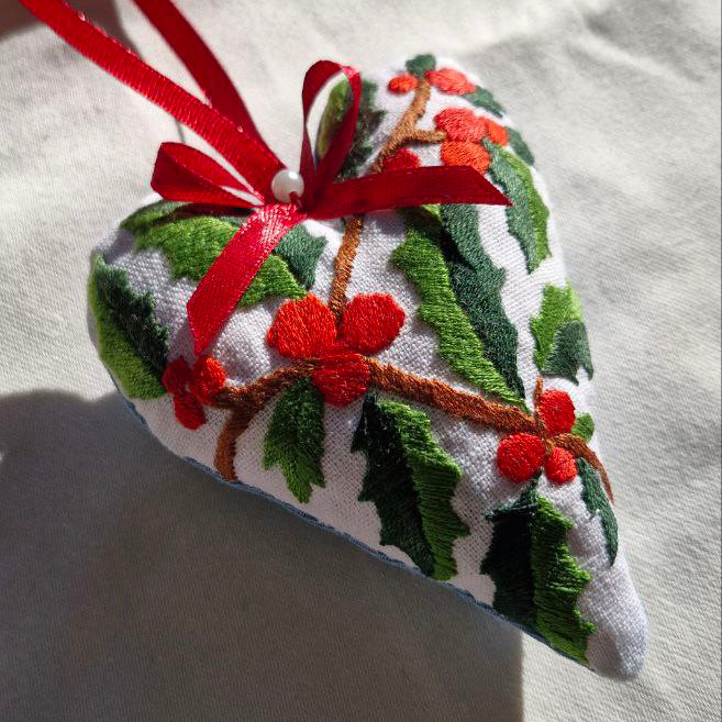 Heart-shaped embroidered ornament with holly design and red ribbon on a white background